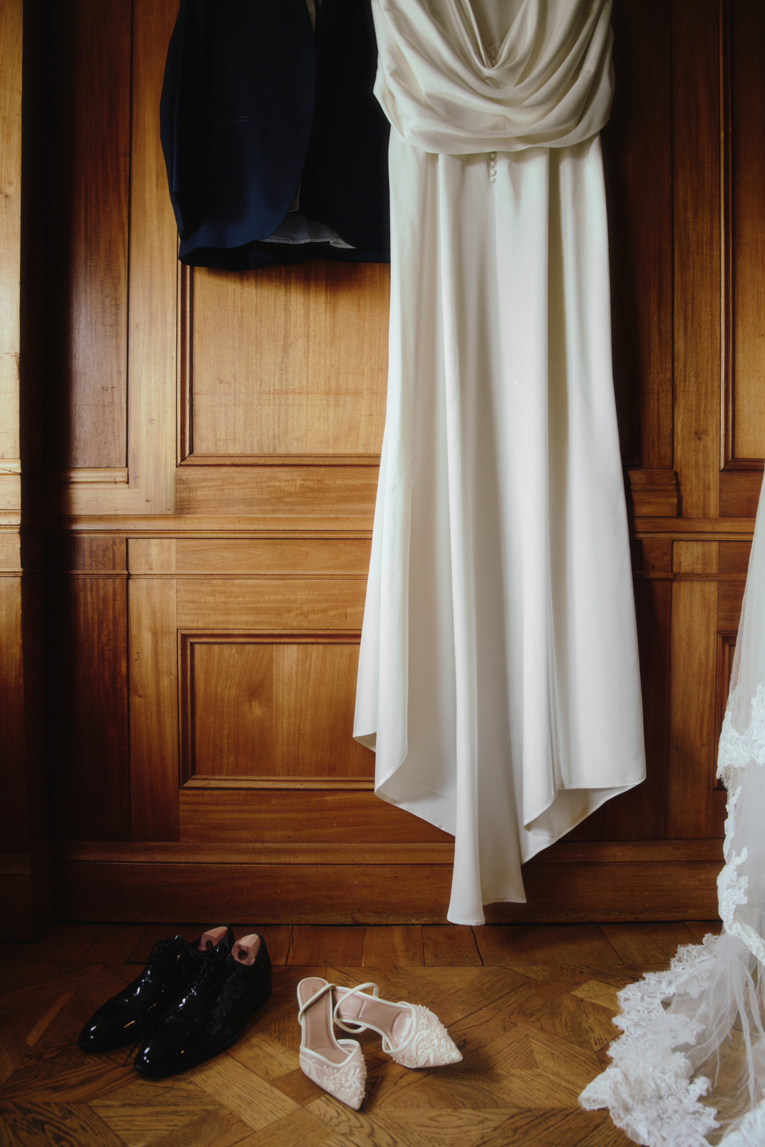 Ivory satin wedding dress hanging against wood panelling, with bridal shoes and a tuxedo nearby.